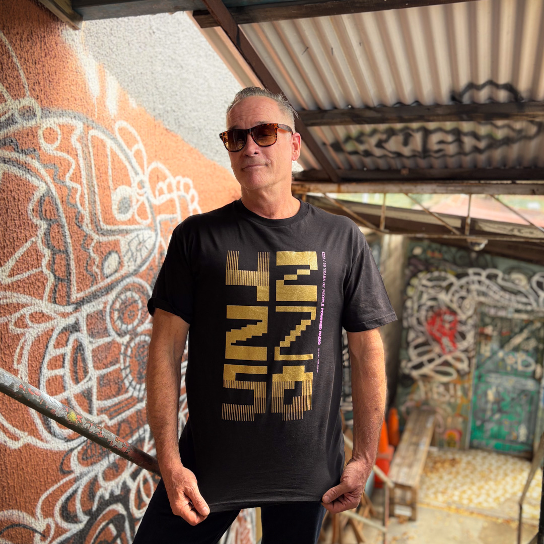 A man wearing sunglasses standing in front of a graffiti covered corner, wearing a black T-shirt featuring the 50th Birthday design