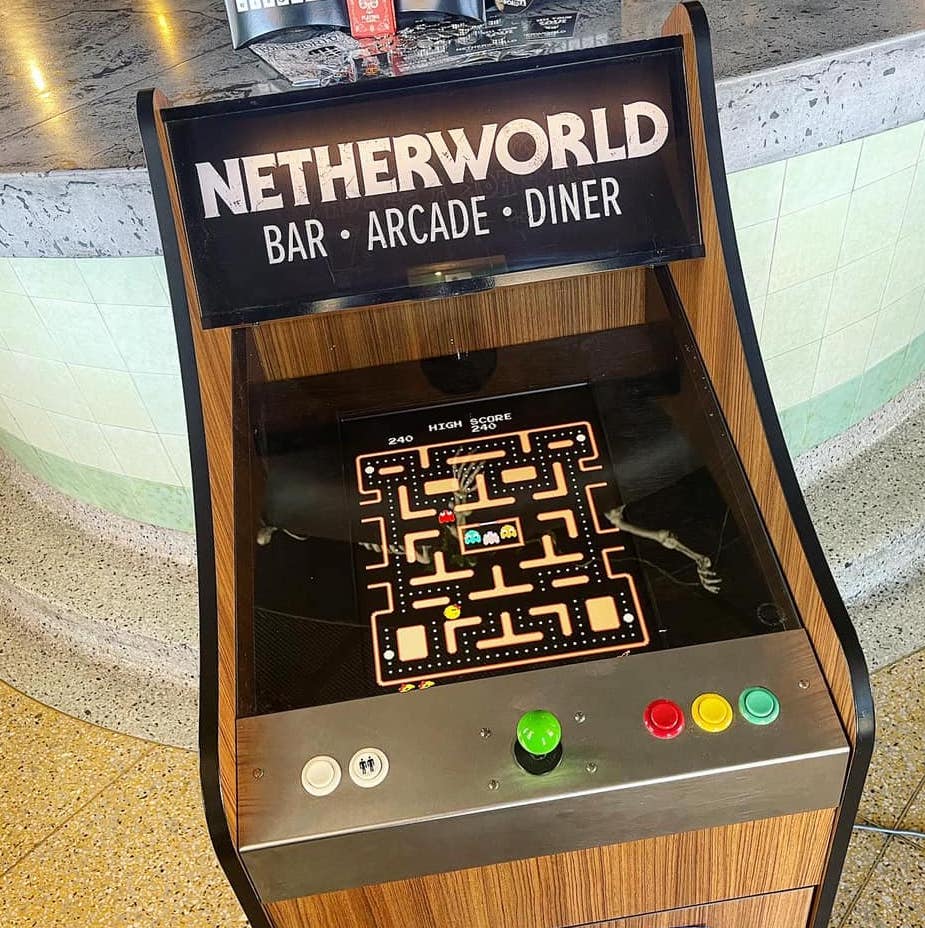 A retro Netherworld arcade game of Pacman in front of the Netherworld bar, with a tiny bone of an arm and claw on top of the screen.