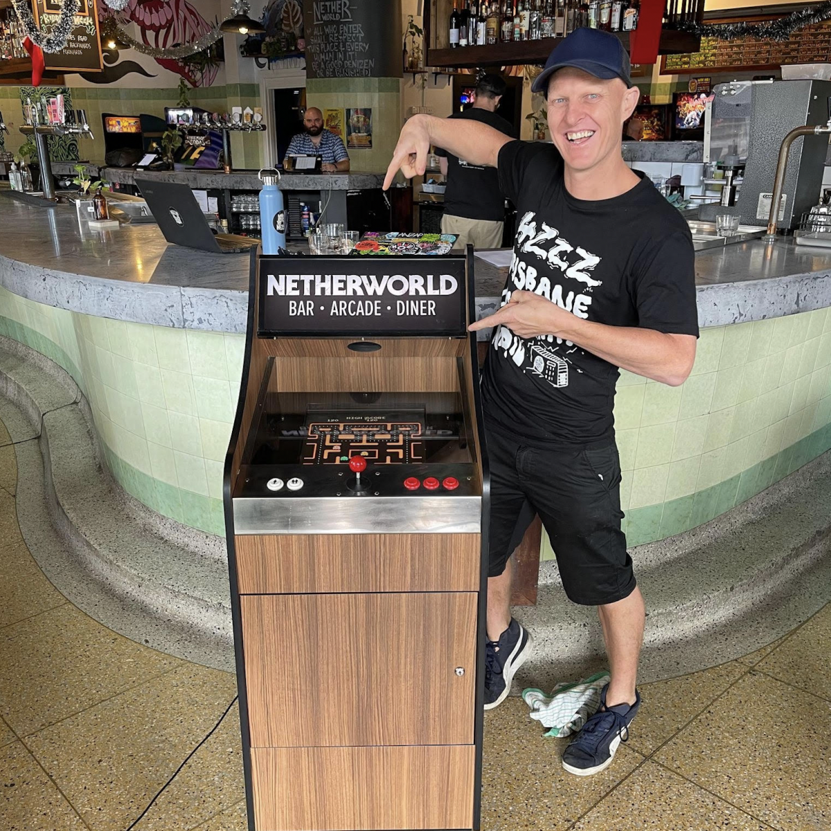 A Netherworld Arcade game cabinet with pacman open on the screen and a person in 4ZZZ merch and a cap in front of the Netherworld bar pointing to it enthusiastically.