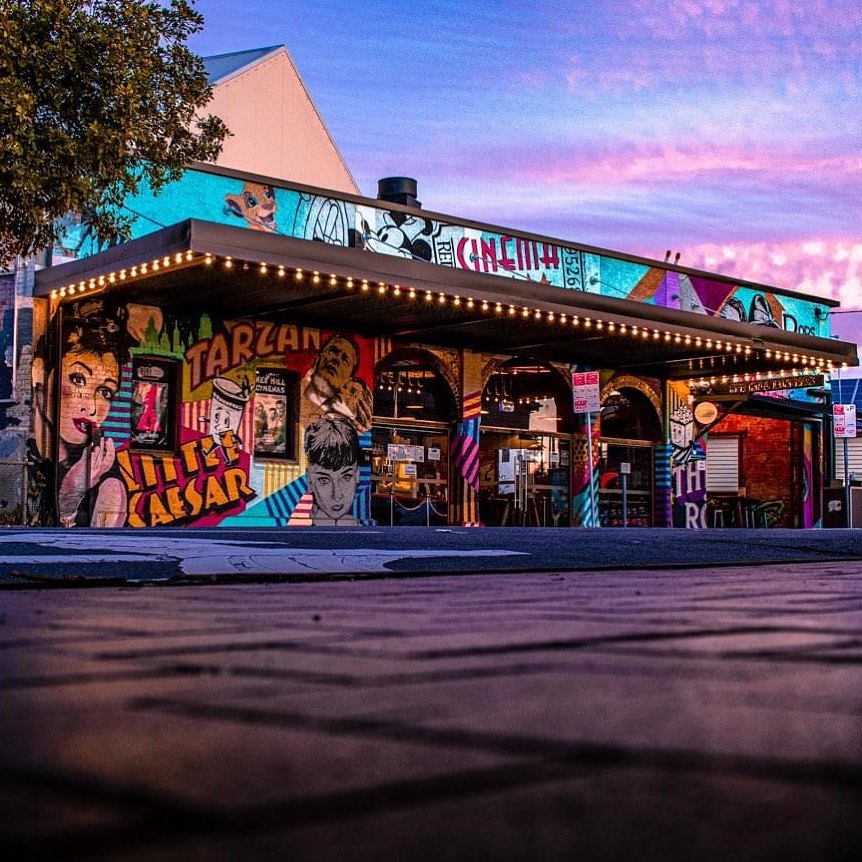 The exterior of Five Star Cinemas Red Hill, painted with colourful retro art of film stars & characters, including Holly Golightly from Breakfast At Tiffany's, Sabrina from the film of the same name, Mickey Mouse, Simba, and Tarzan and Jane. In the foreground, the road is glistening from recent rain and, in the background, the cumulo-stratus clouds are reflecting beautiful blue & purple hues.