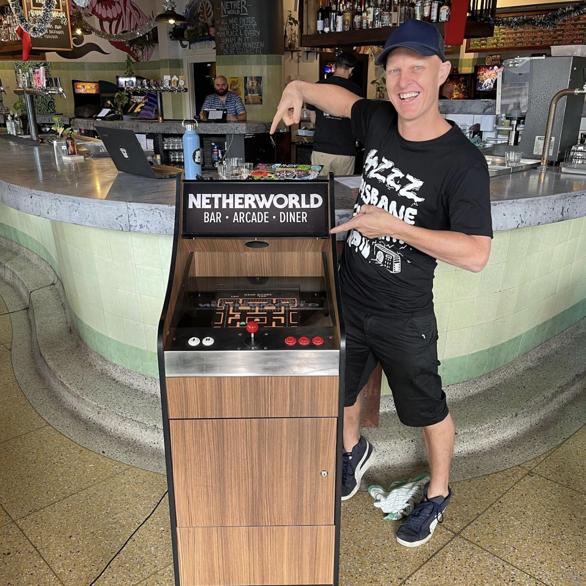 Jimmy, one of the head honchos at Netherworld is standing beside a lowboy arcade cabinet wearing a black t-shirt with a design that reads '4ZZZ Brisbane Community Radio', black shorts & a black cap. Jimmy's smiling & pointing at the arcade cabinet, which one lucky 4ZZZ subscriber will win this Radiothon!