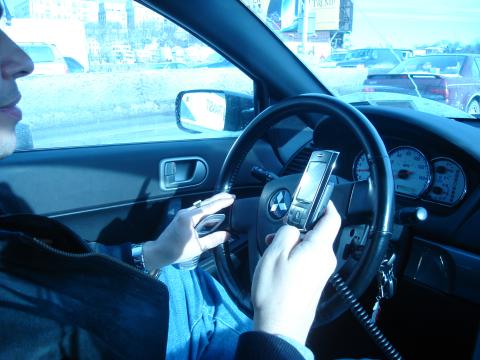 A driver with their left hand on the wheel and their right hand using their mobile phone.